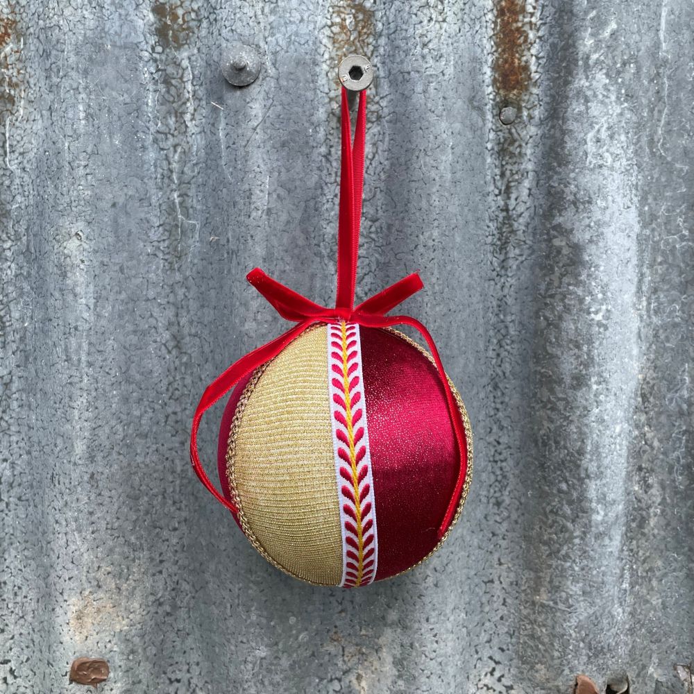 Velvet round bauble in red and gold fabric with gold and patterned braiding and red velvet bow on corrugated iron wall