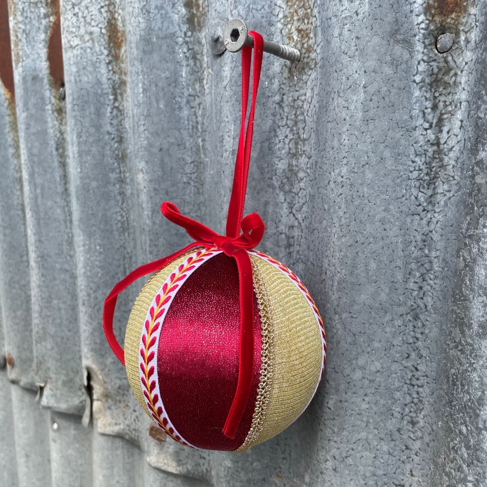 Velvet round bauble in red and gold fabric with gold and patterned braiding and red velvet bow on corrugated iron wall