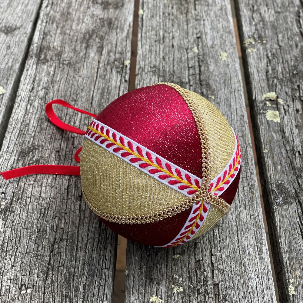 Velvet round bauble in red and gold fabric with gold and patterned braiding and red velvet bow on wooden table