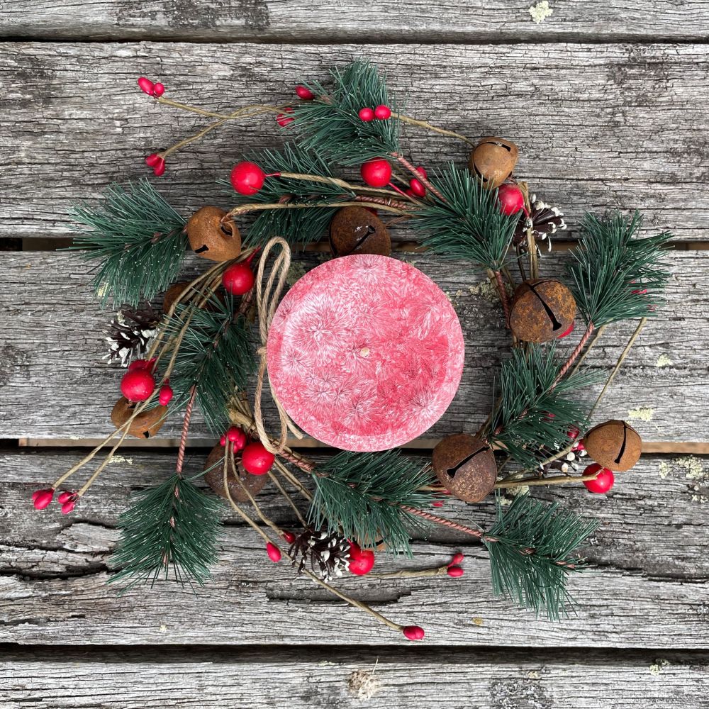 Red palm wax candle on a decorative wreath with greenery, berries, and pinecones on a wooden surface.