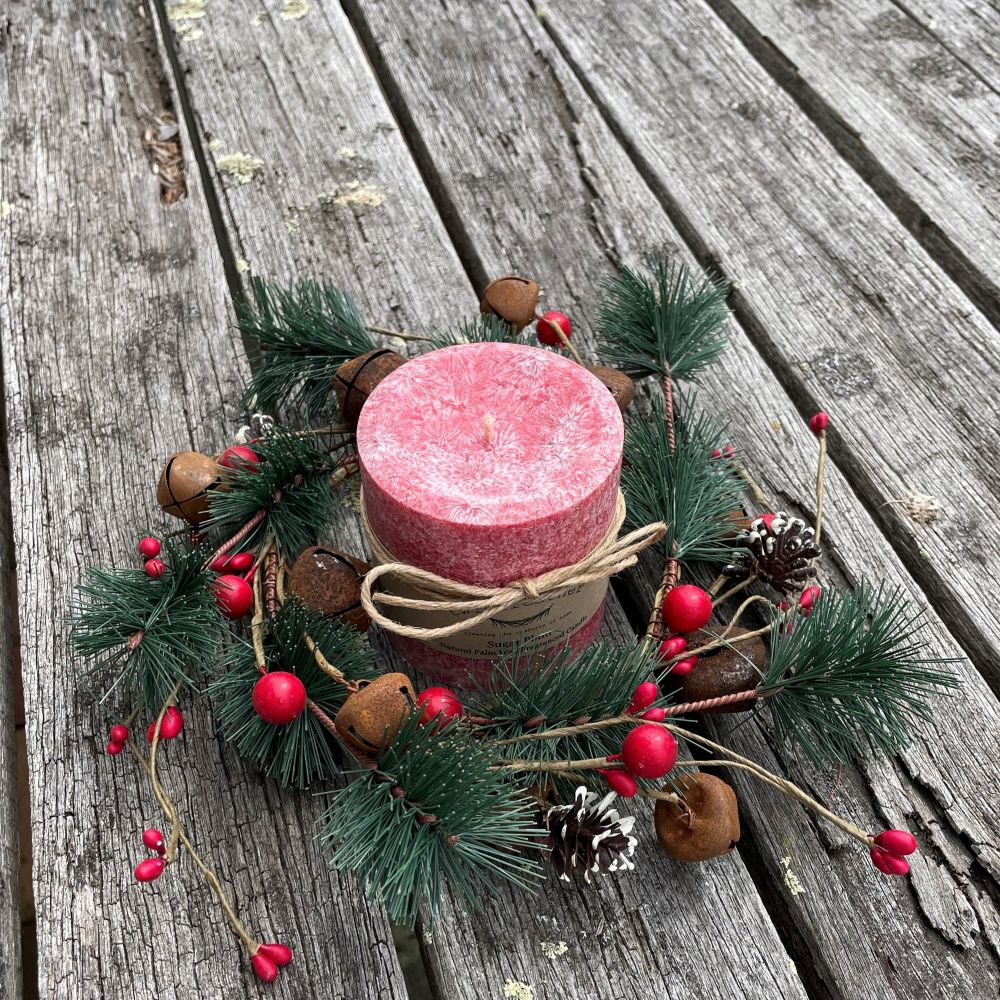 Red palm wax candle on a decorative wreath with greenery, berries, and pinecones on a wooden surface.