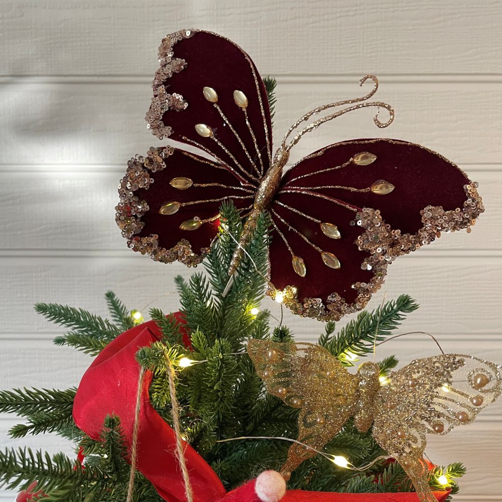 Decorative velvet butterfly in burgundy on the top of a Christmas tree with lights and red ribbon.