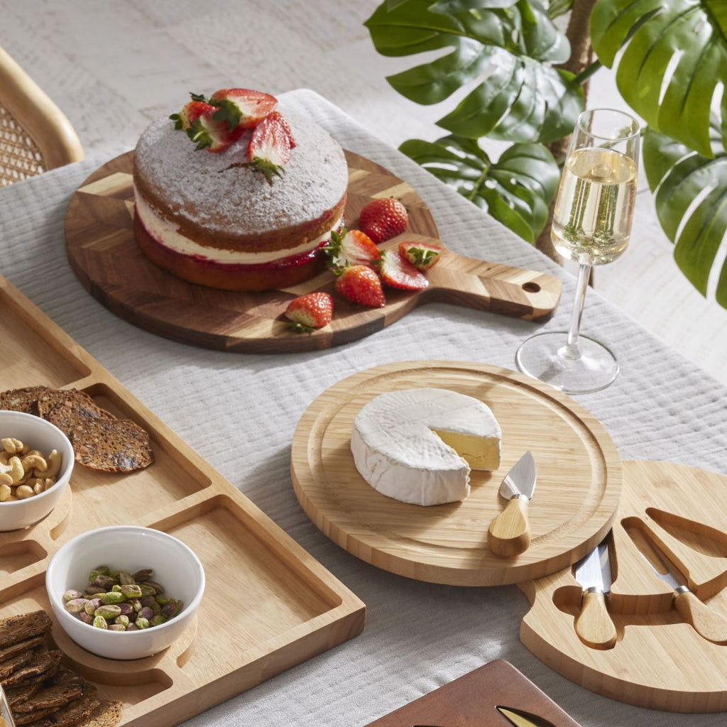 Table setting with a cake on the herringbone cutting board, cheese, and wine on a white tablecloth with greenery in the background.