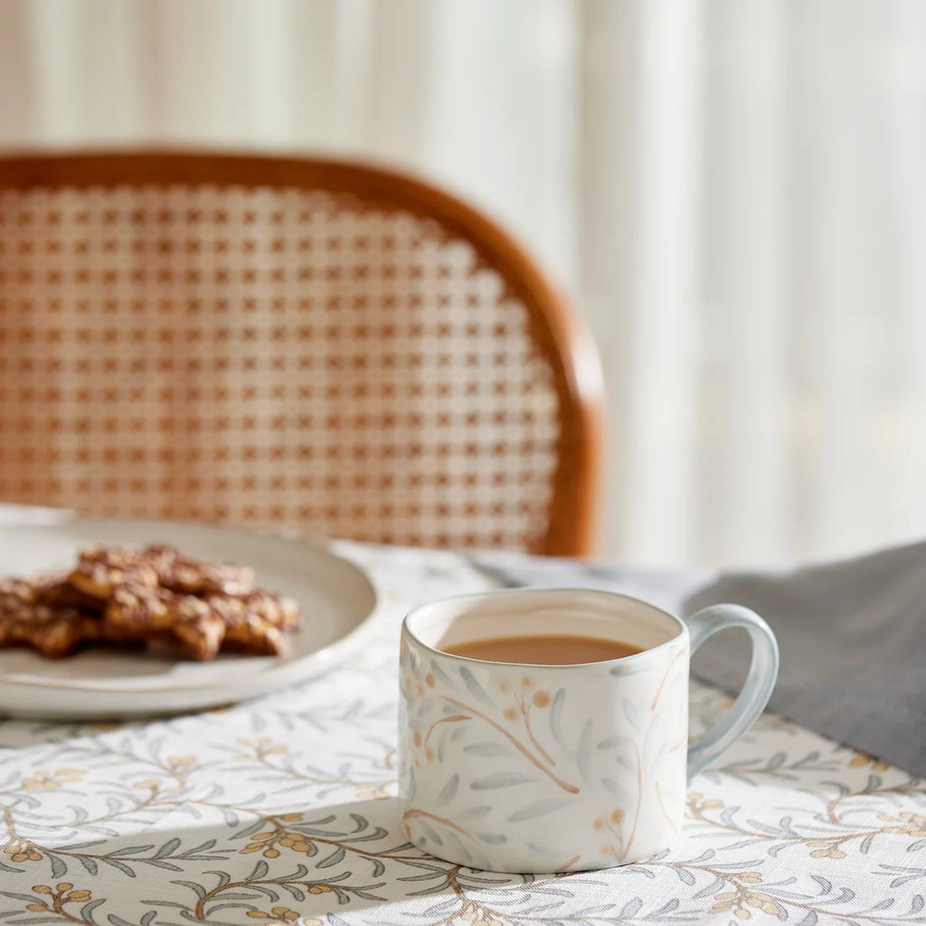 Cup of coffee in a Juniper mug on a patterned Juniper tablecloth with cookies and a chair in the background