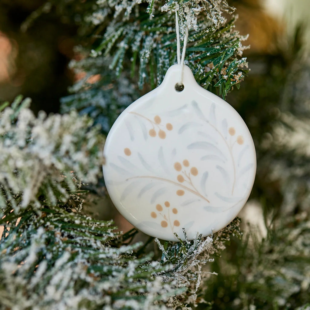 Round Juniper hanging white ornament with subtle design of blue leaves and apricot berries hanging on a frosted Christmas tree.
