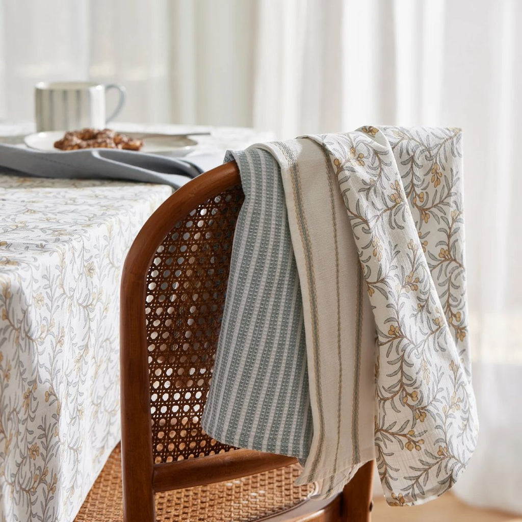 Dining table with a juniper floral tablecloth, wicker chair, and three juniper tea towels hanging on back of chair.