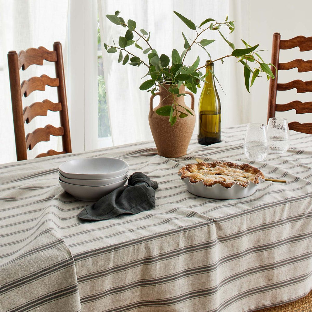 Dining table with a charcoal striped tablecloth, pie, bowls, and a vase with greenery.