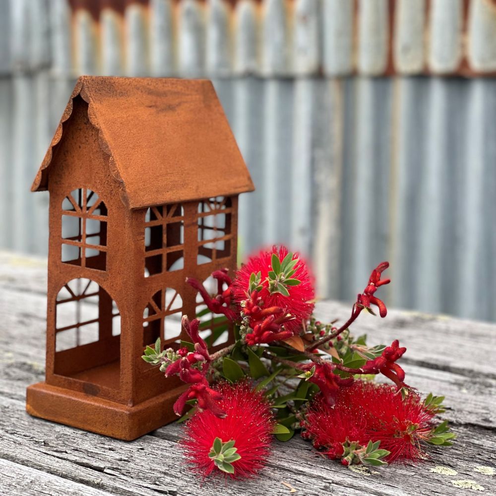 Large rusty metal house shaped lantern or candle holder on a wooden table with an arrangement of Australian native flowers next to it