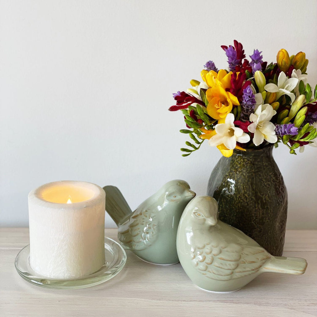 Decorative items including a lit Twig & Feather Gardenia candle, ceramic birds, and a green organic shaped vase with freesias on a table.