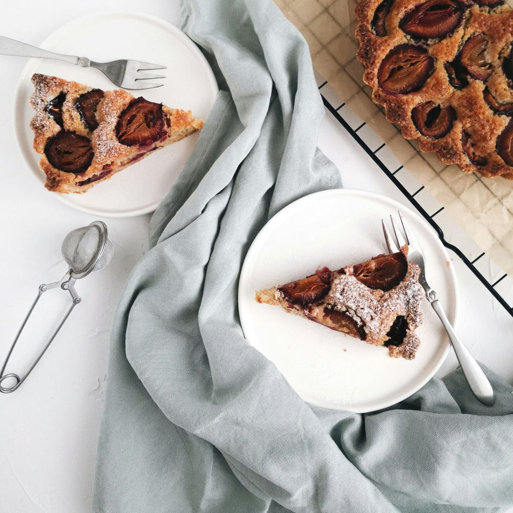 Two slices of plum tart on white plates with a whole tart in the background, on a light blue cloth.