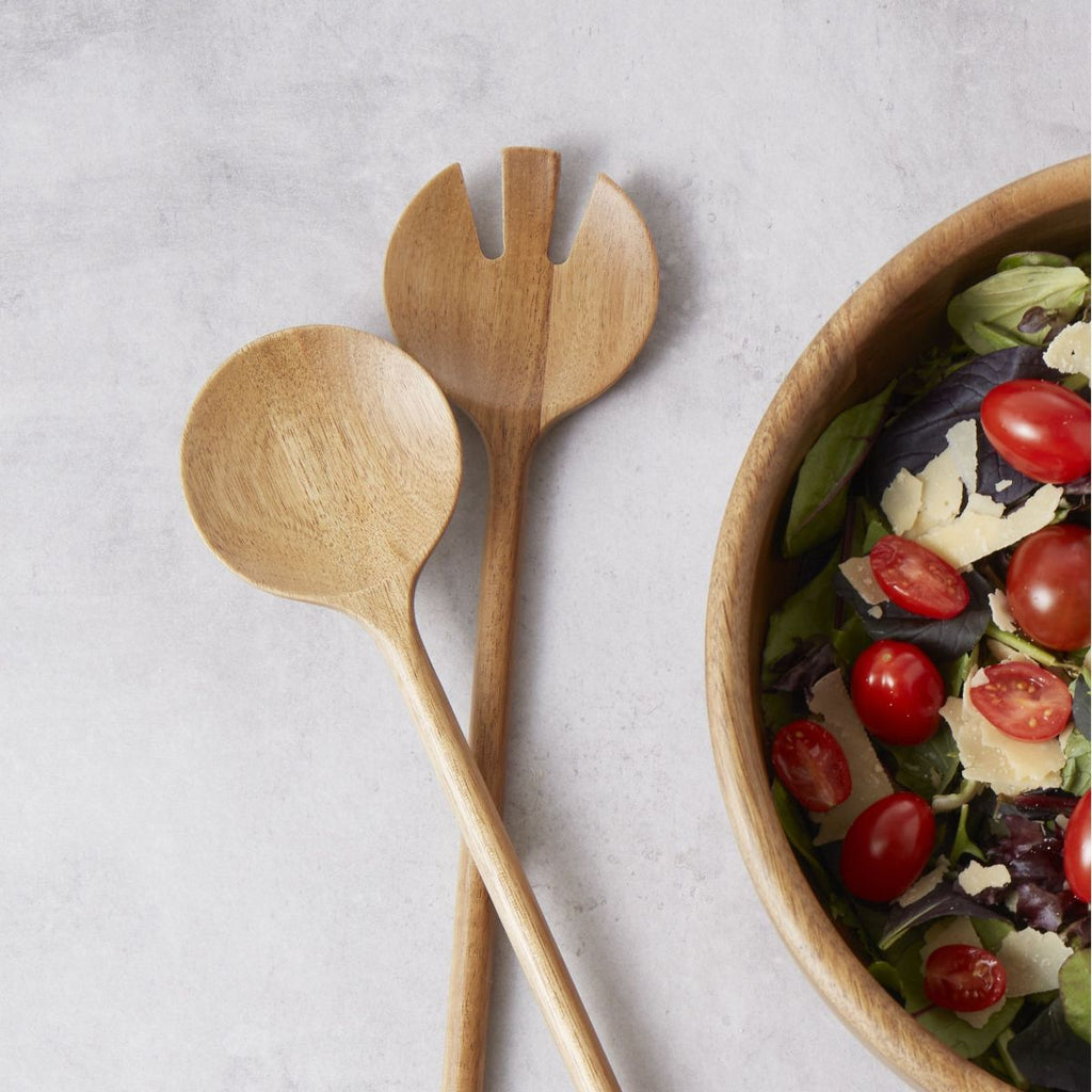 Acacia wood salad servers next to a bowl of salad on a light gray background
