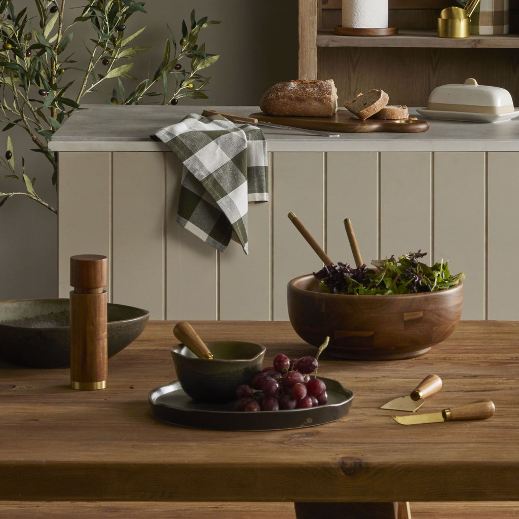 Kitchen scene with Acacia wood salad servers, wooden table, bowls, and a checkered towel.
