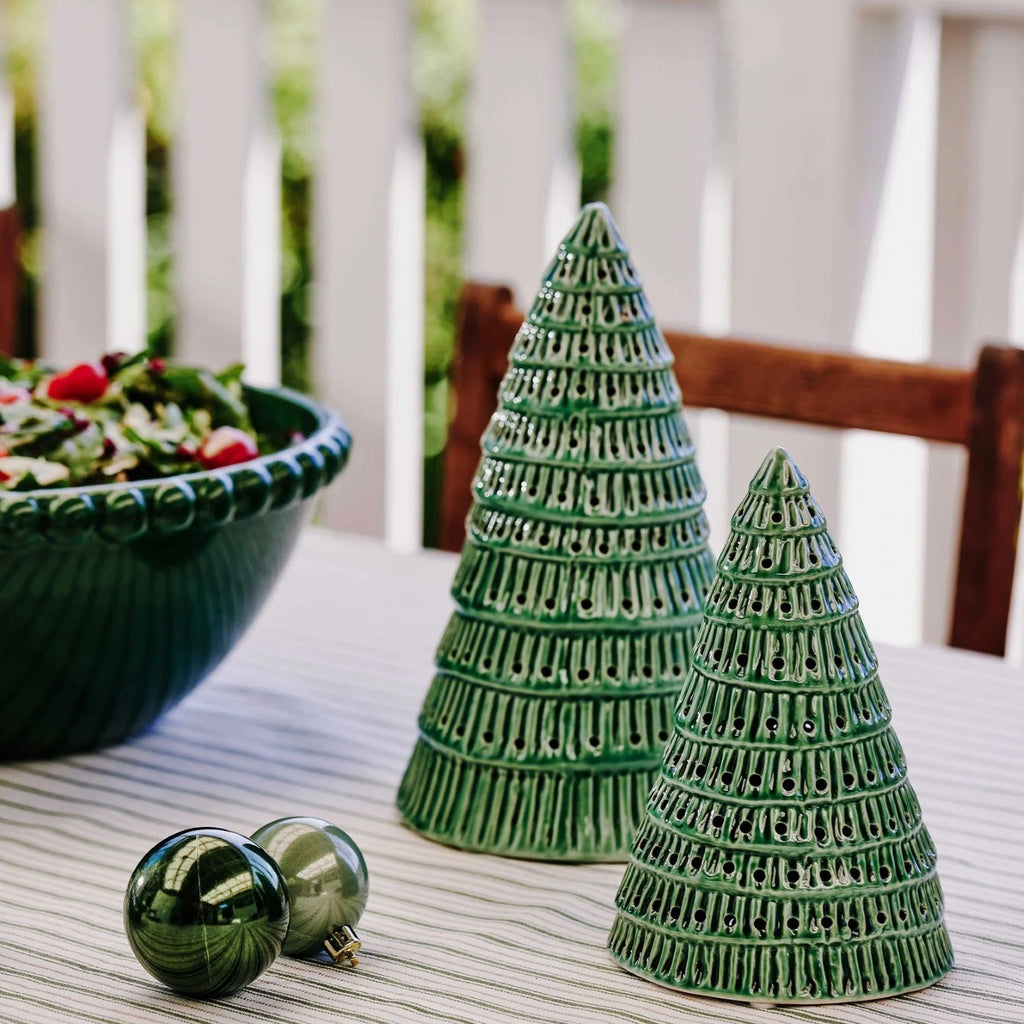 Two green ceramic Christmas trees on a table with a bowl of fruit in the background.