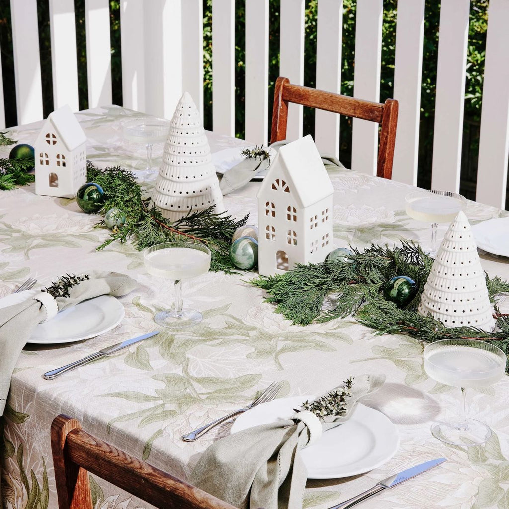 Decorative Christmas table setting with white ceramic houses, White ceramic Christmas trees, greenery, and ornaments on a white tablecloth.