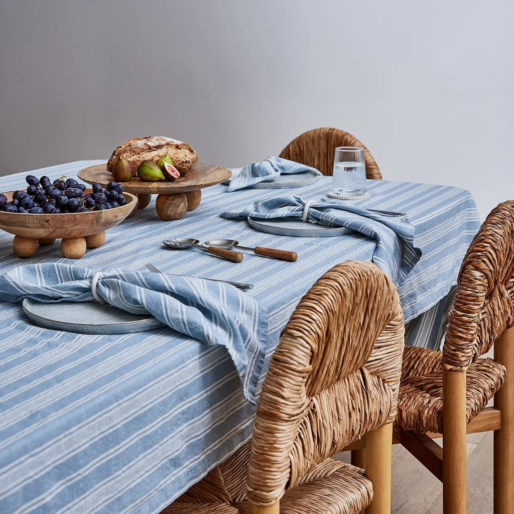 Dining table set with a blue striped tablecloth and napkins, wicker chairs, and a small meal