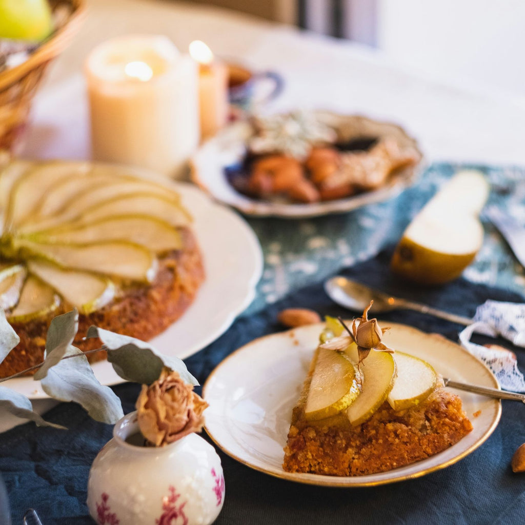 Slice of cake with pear on a plate, surrounded by a cake and candles on a table - evokes the feeling of the pear and ginger fragrance