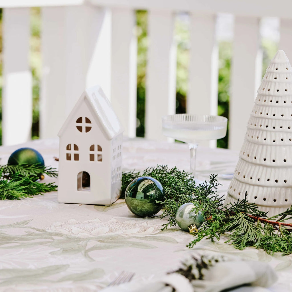 Decorative table setting with white ceramic house, tree, and green ornaments on a white surface.