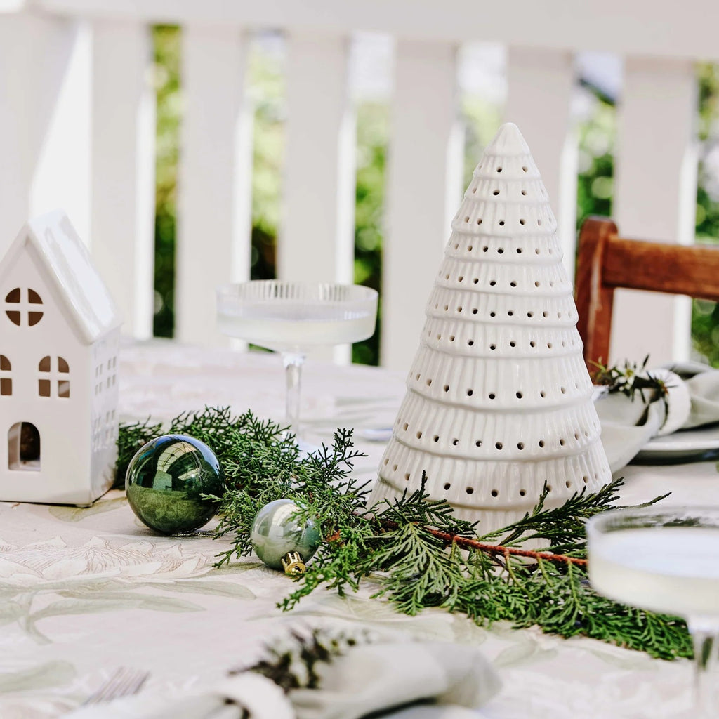 Decorative table setting with white ceramic Christmas tree, house, and greenery on a white tablecloth.