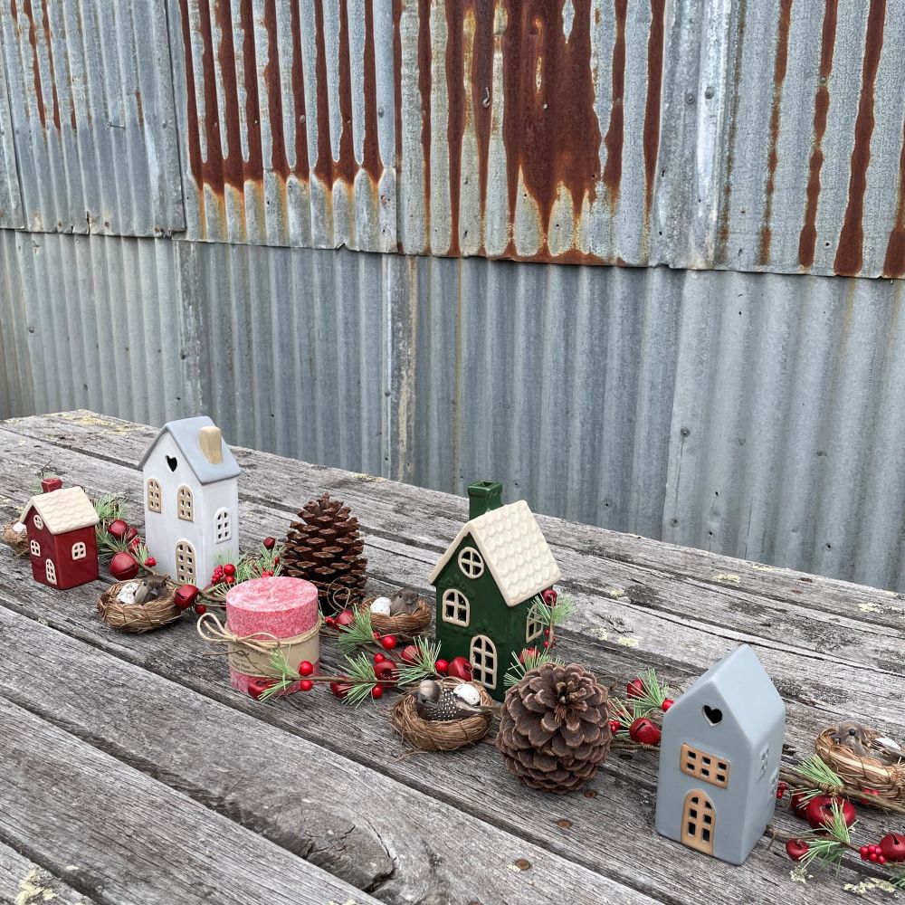 Decorative miniature houses, a red bell garland and pinecones on a wooden surface with a corrugated metal background.