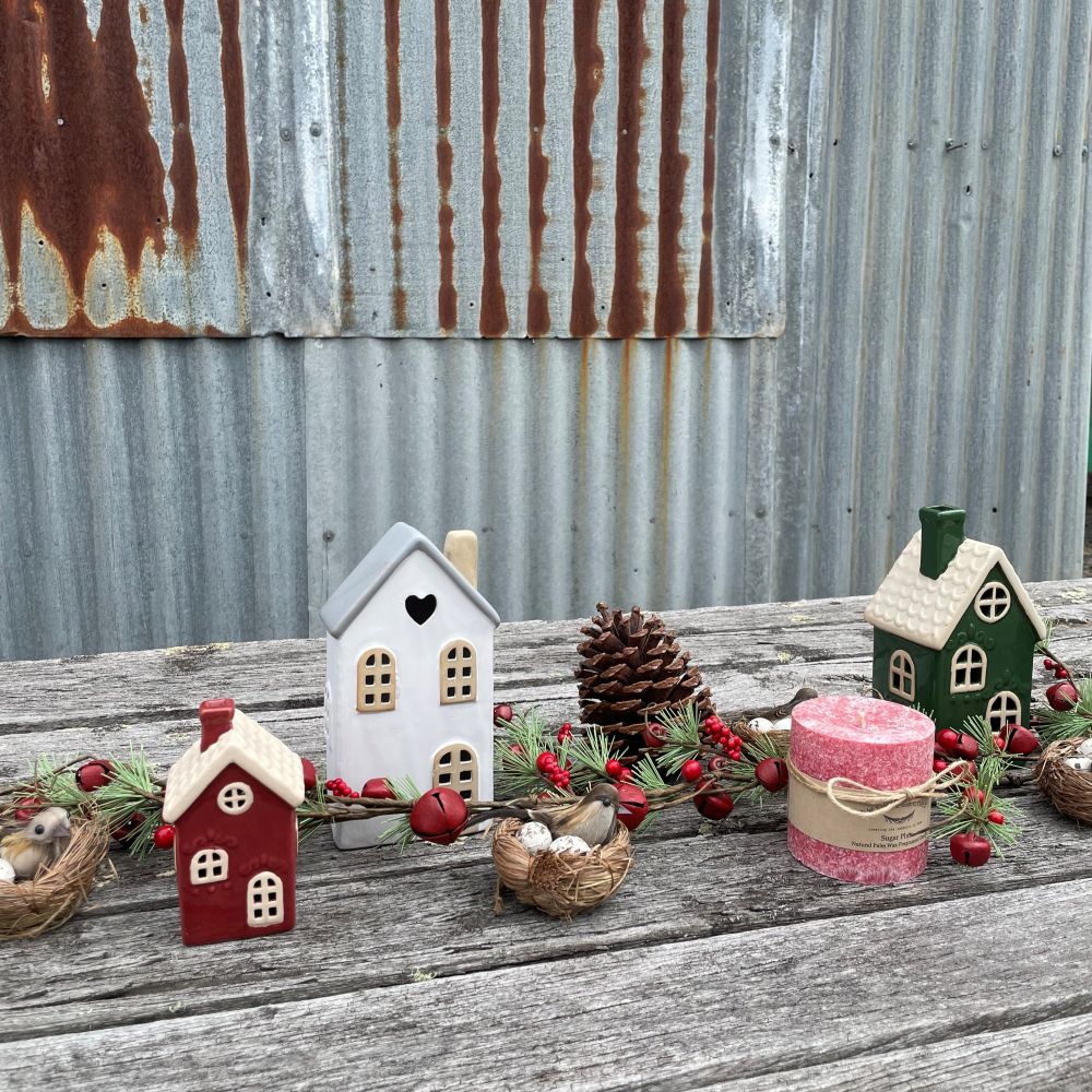 Decorative setup with small houses, candles, and pinecones on a rustic wooden surface against a corrugated metal wall.