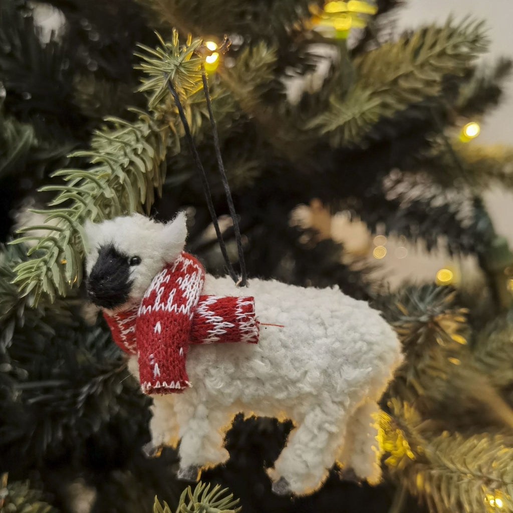 White sheep ornament with a red sweater hanging on a decorated Christmas tree.