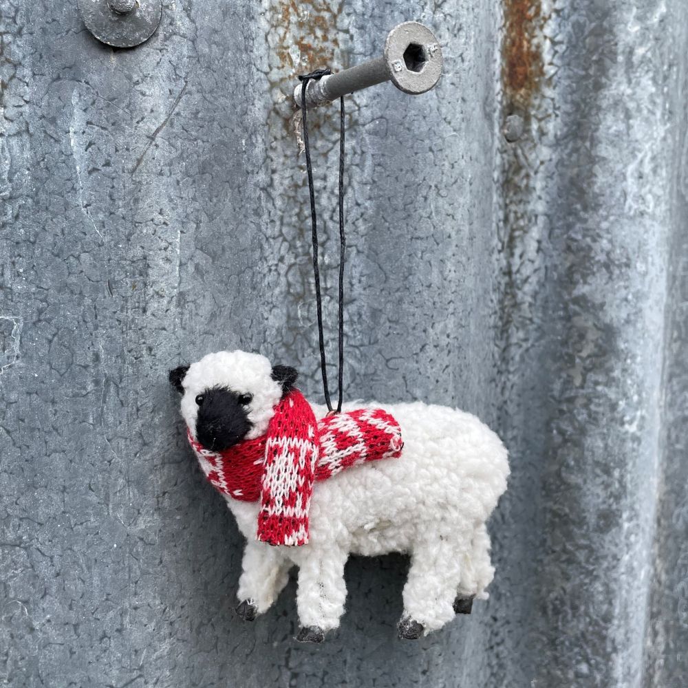White sheep ornament with a red and white patterned scarf hanging on a textured metal surface.
