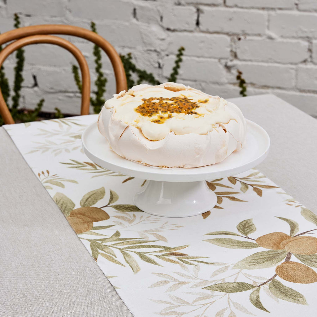 White pavlova dessert on a white cake stand with a Spring Orchard table runner on a table.