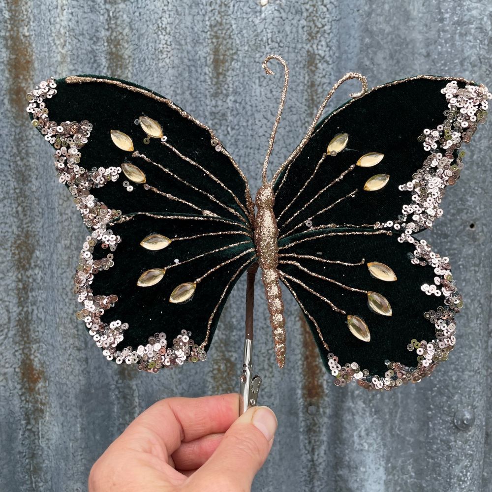 Decorative butterfly with dark green wings and gold accents held by a hand against a textured gray background.