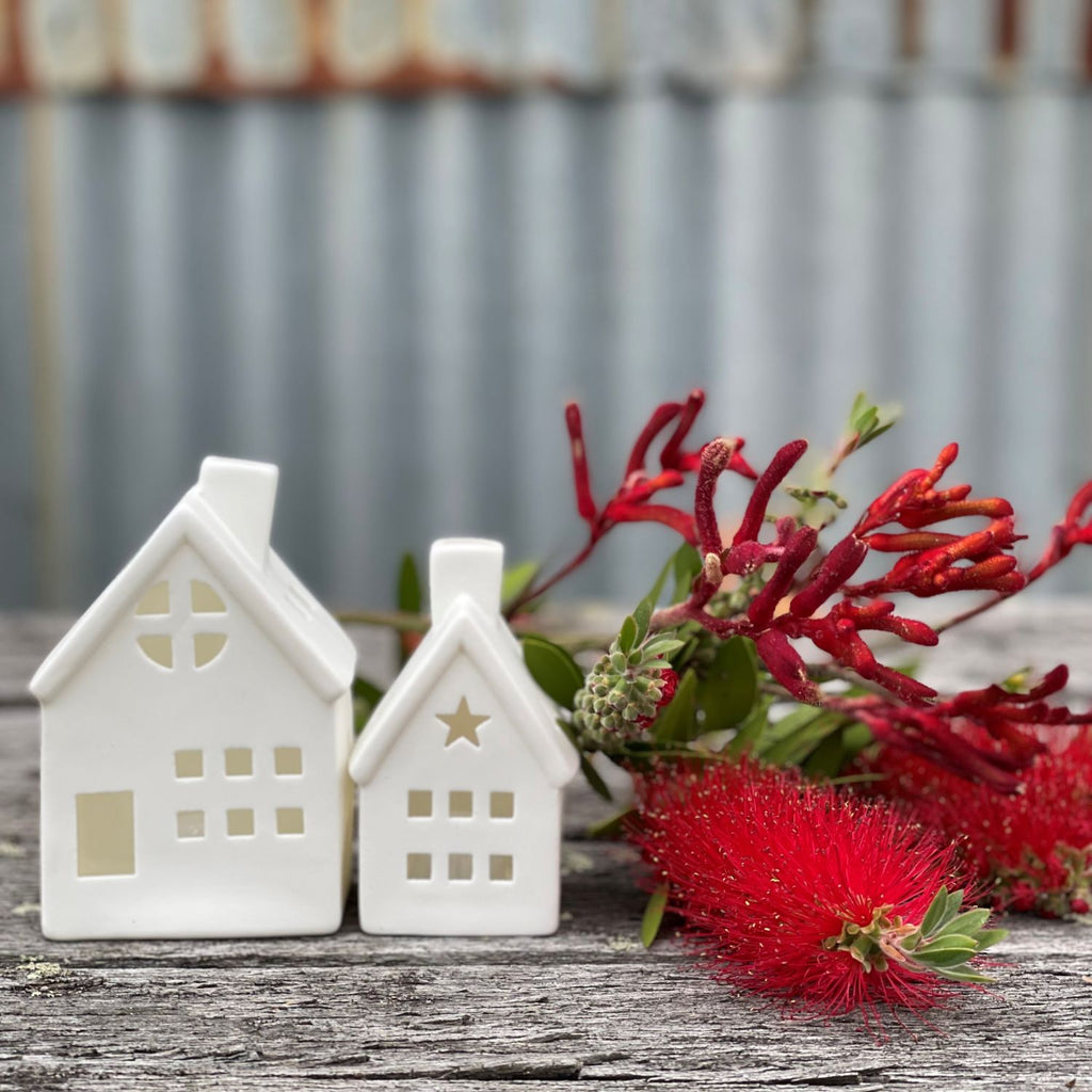 White porcelain houses in x-small and small with chimney, windows and star cut out with LED light sitting on a wooden table with Australian native flowers next to them