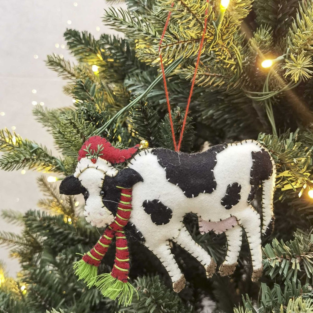 Felt Cow Christmas ornament with a red hat and scarf hanging on a decorated tree.