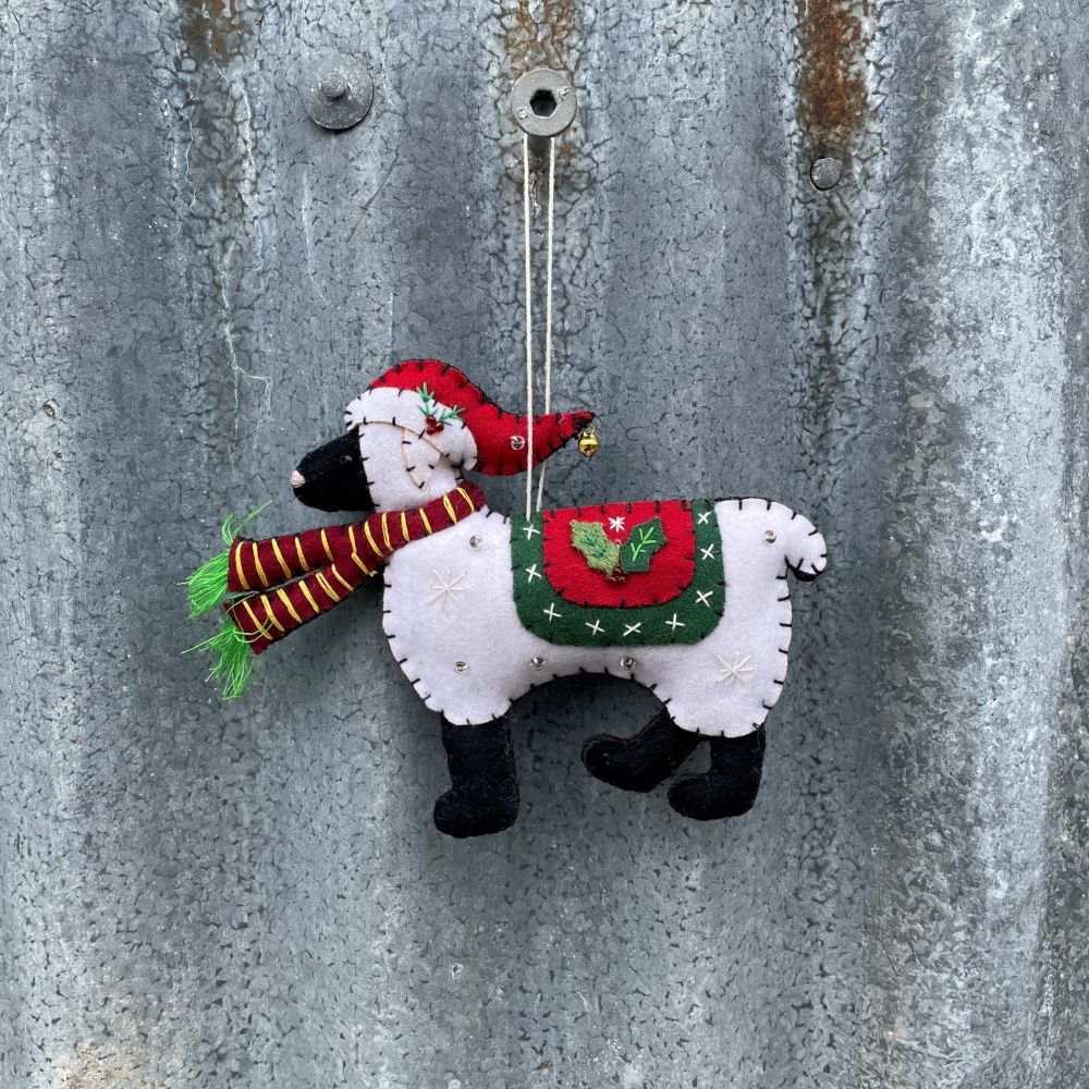 Felt ornament of a sheep with a red hat and green bag hanging on a textured metal surface.