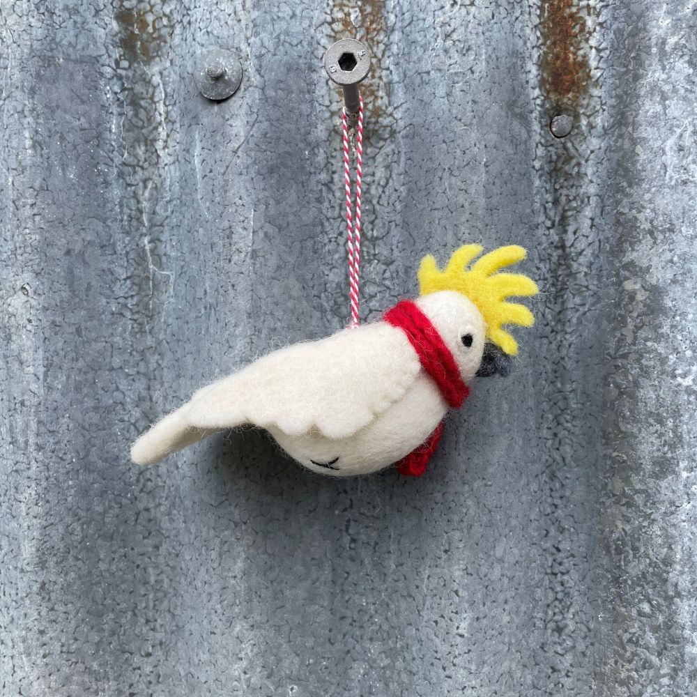 Felt cockatoo bird ornament with a red scarf and yellow crest hanging on a textured metal surface.