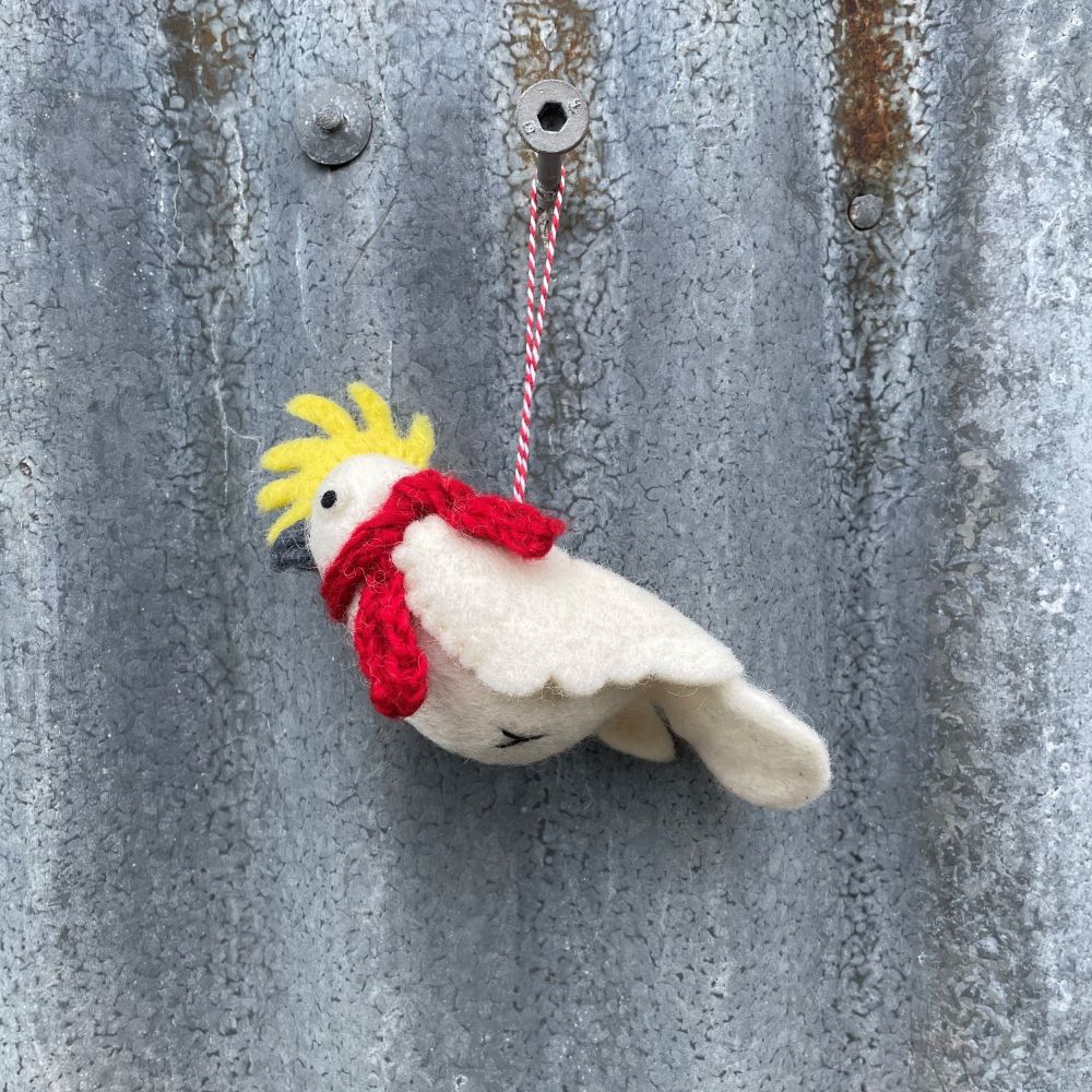Felt cockatoo bird ornament with a red scarf and yellow crest hanging on a textured metal surface.