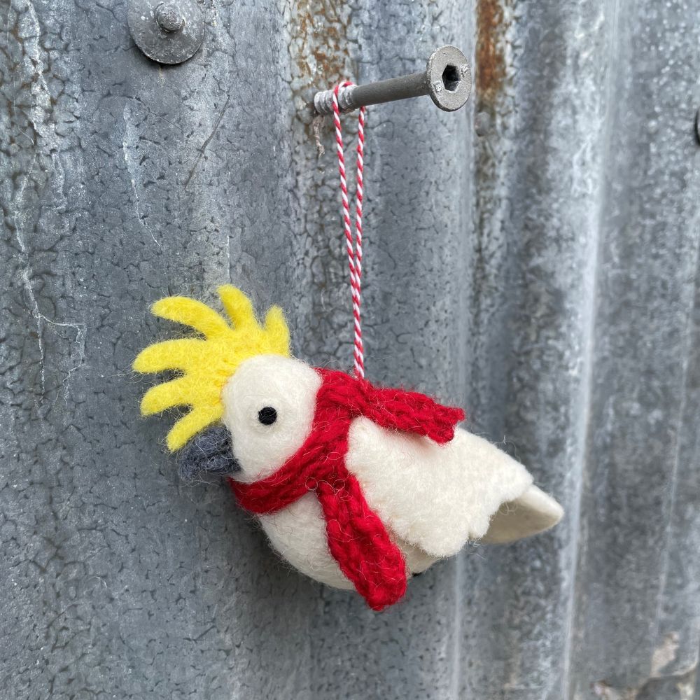 Felt cockatoo bird ornament with a red scarf and yellow crest hanging on a textured metal surface.
