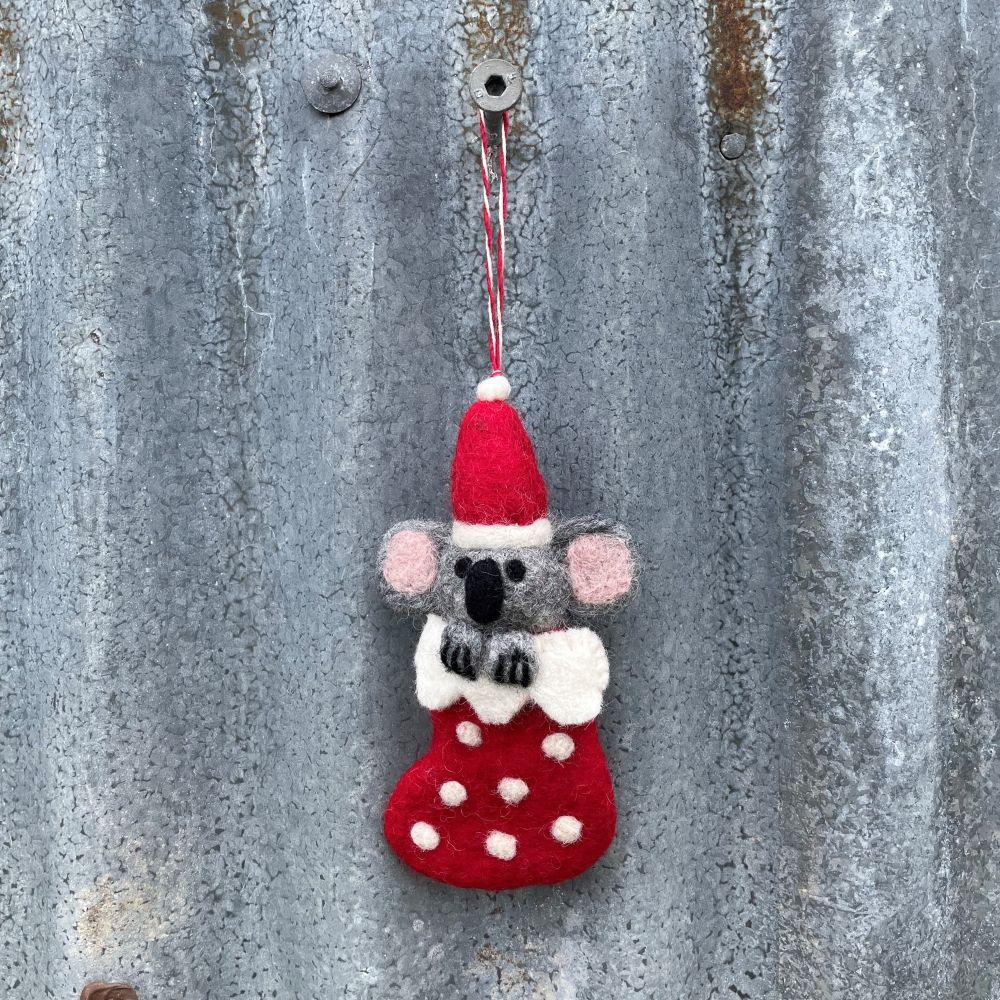 Felted koala ornament in a red stocking with a Santa hat on a metal surface