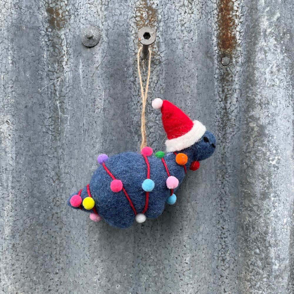 Felted dinosaur ornament in dark blue with a Santa hat and Christmas lights on a textured metal surface