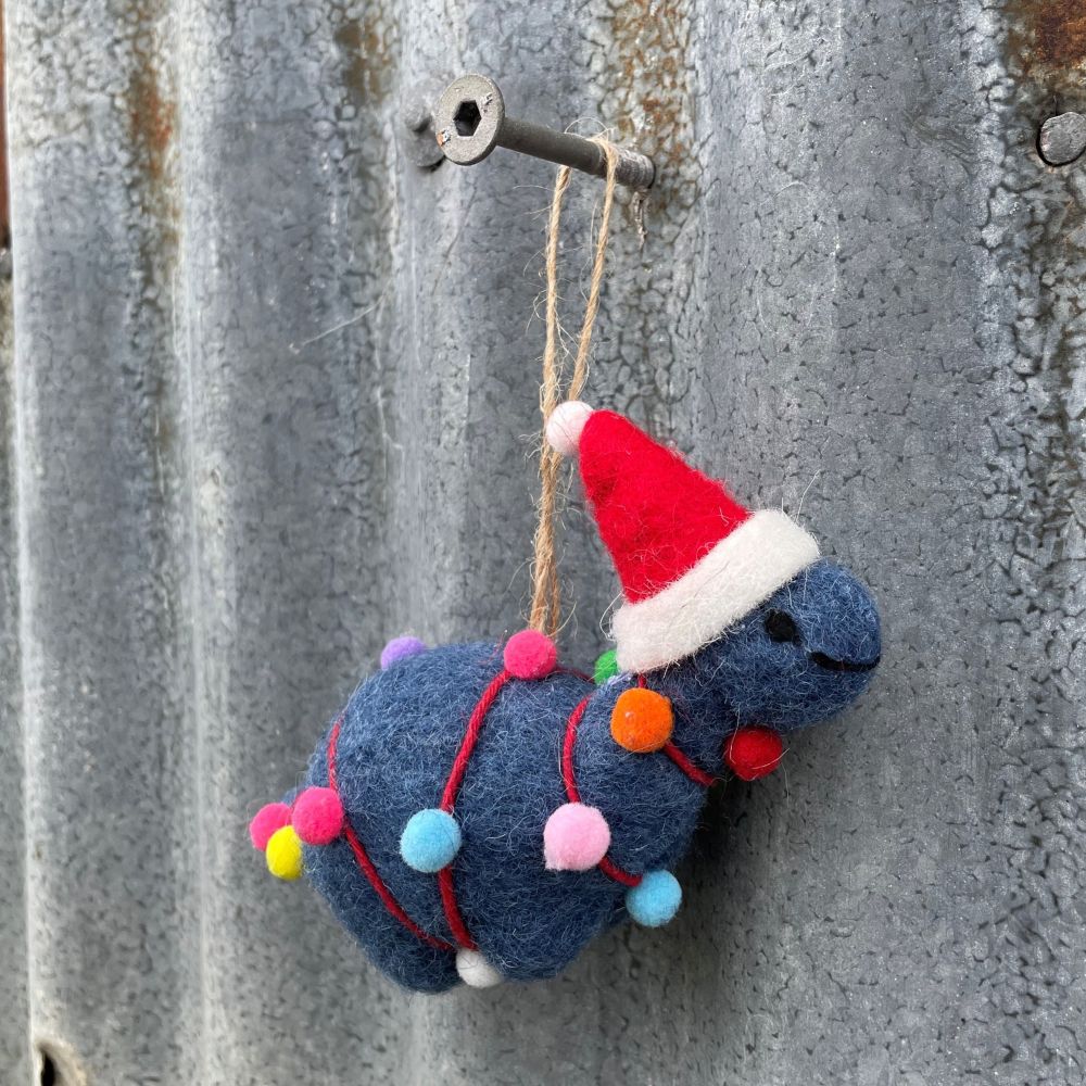 Felted dinosaur ornament in dark blue with a Santa hat and Christmas lights on a textured metal surface