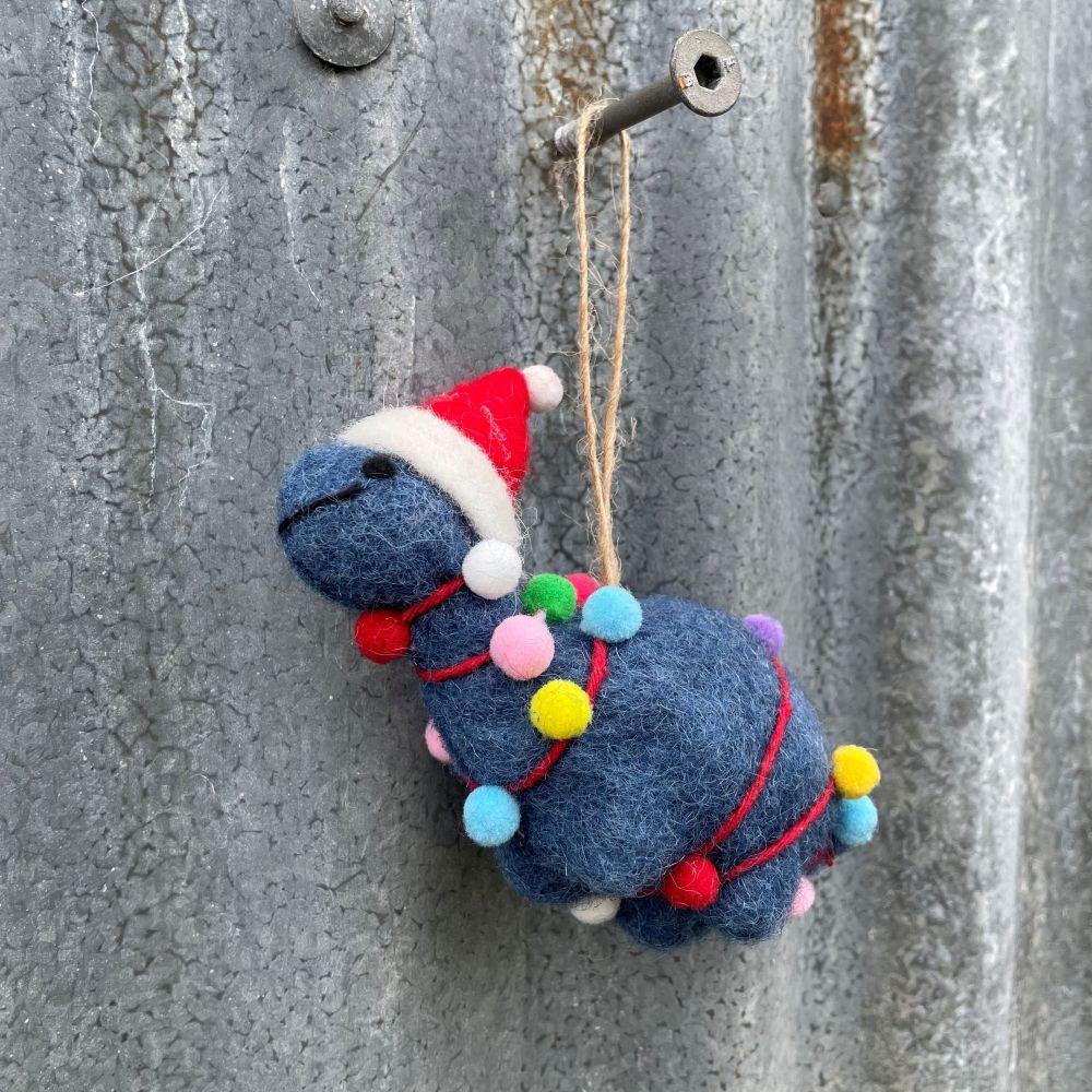 Felted dinosaur ornament in dark blue with a Santa hat and Christmas lights on a textured metal surface