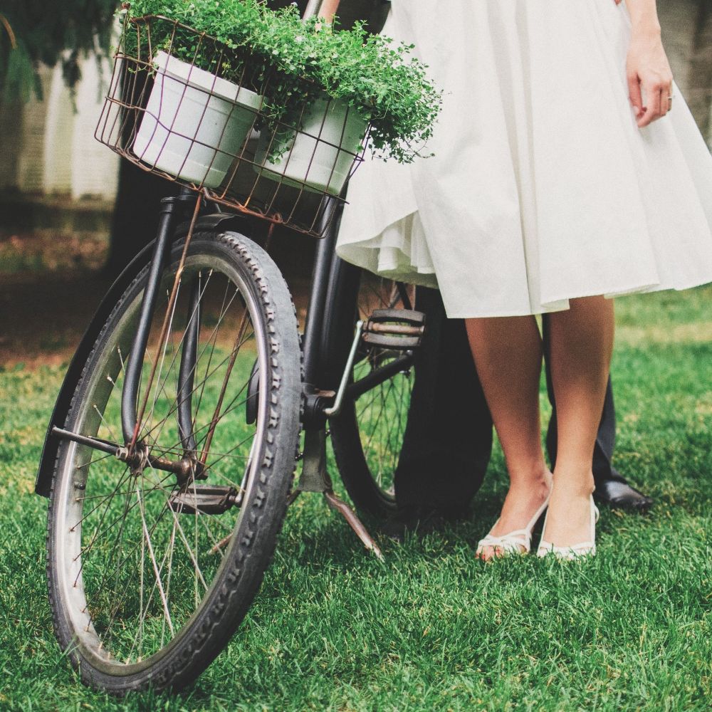 Person in a white dress standing next to a bicycle with a basket of plants on grass