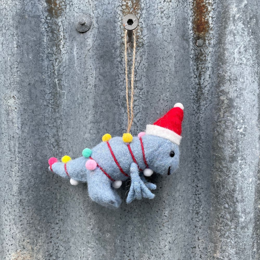 Felted dinosaur ornament in light blue with a Santa hat and Christmas lights on a textured metal surface