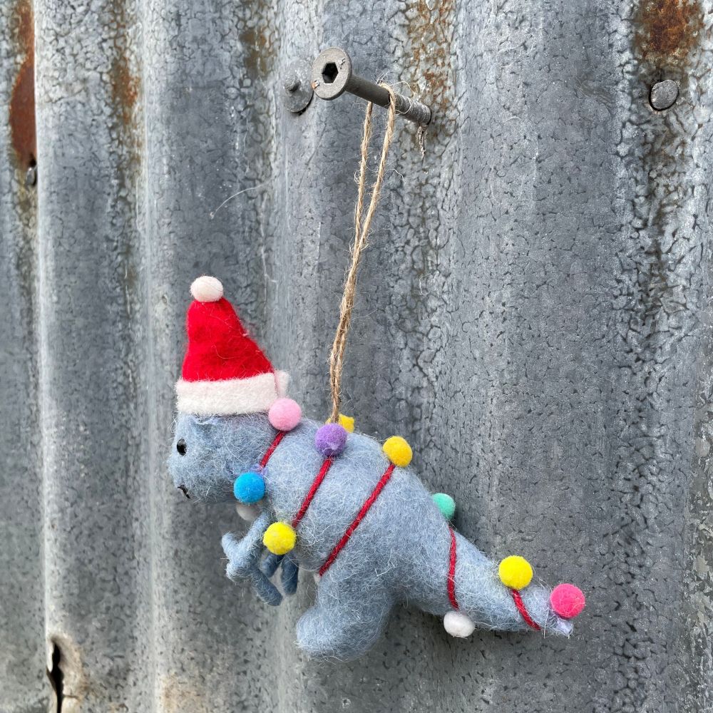 Felted dinosaur ornament in light blue with a Santa hat and Christmas lights on a textured metal surface