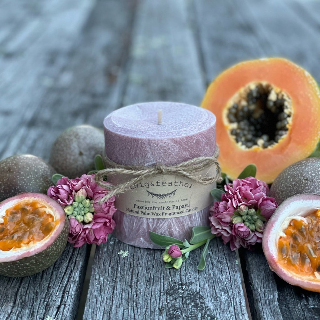 Twig and Feather Passionfruit and Papaya pillar 38 hour palm wax candle surrounded by passionfruit halves, papaya, and flowers on a wooden surface.