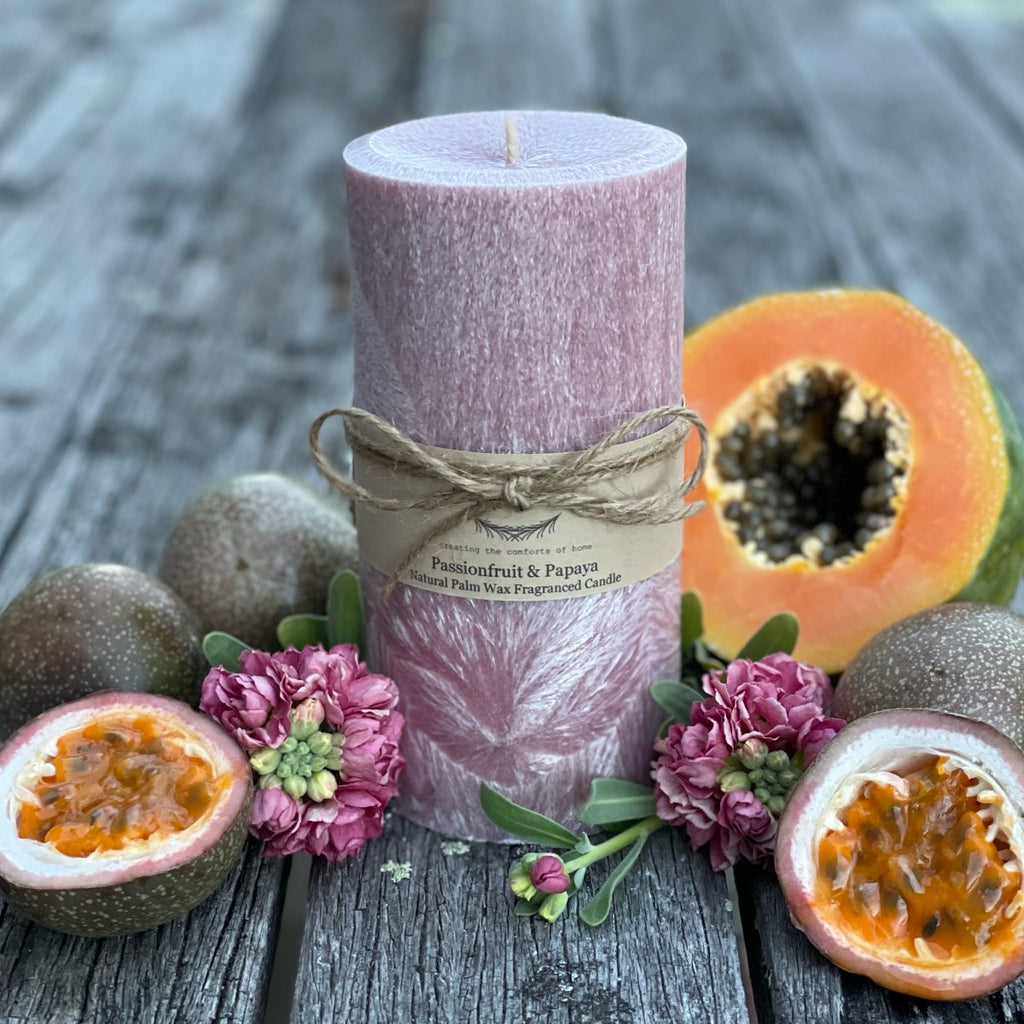 Twig and Feather Passionfruit and papaya palm wax pillar candle 80hrs- Mauve candle surrounded by halved passionfruit, papaya, and flowers on a wooden surface.