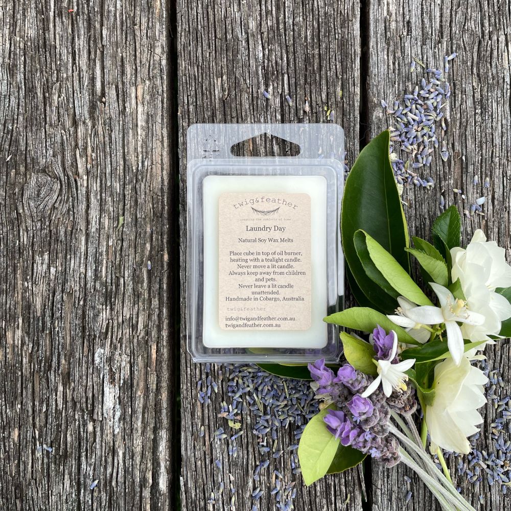 Twig and Feather Laundry Day scented wax melt in a clear container on a wooden surface with lavender, blossoms and green leaves.